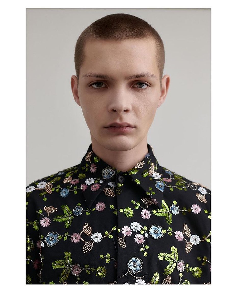 Close-up of an adult male wearing a black button-down shirt with multicolor floral embroidery, posing in front of a neutral background.