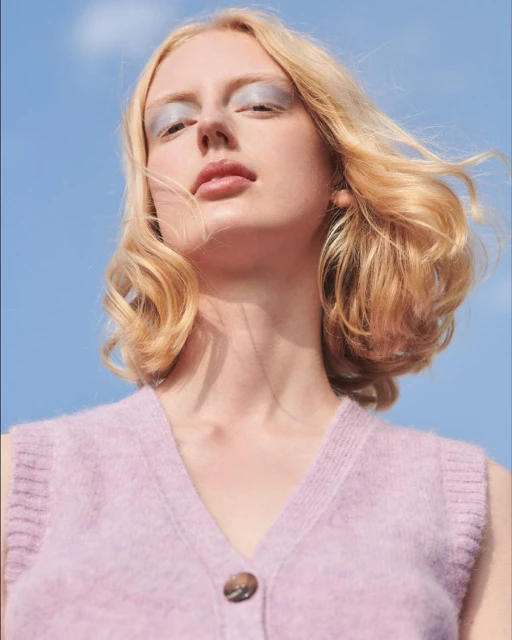 Blonde woman with blue eyeshadow and a lavender sweater vest, poses against a blue sky in a beauty or fashion shot.