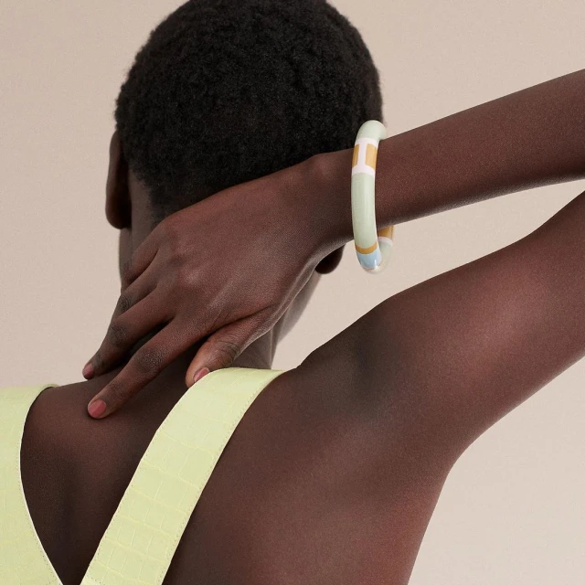 An unrecognizable person with dark skin tones is wearing a pastel-colored resin bangle, photographed from the back in a studio setting.