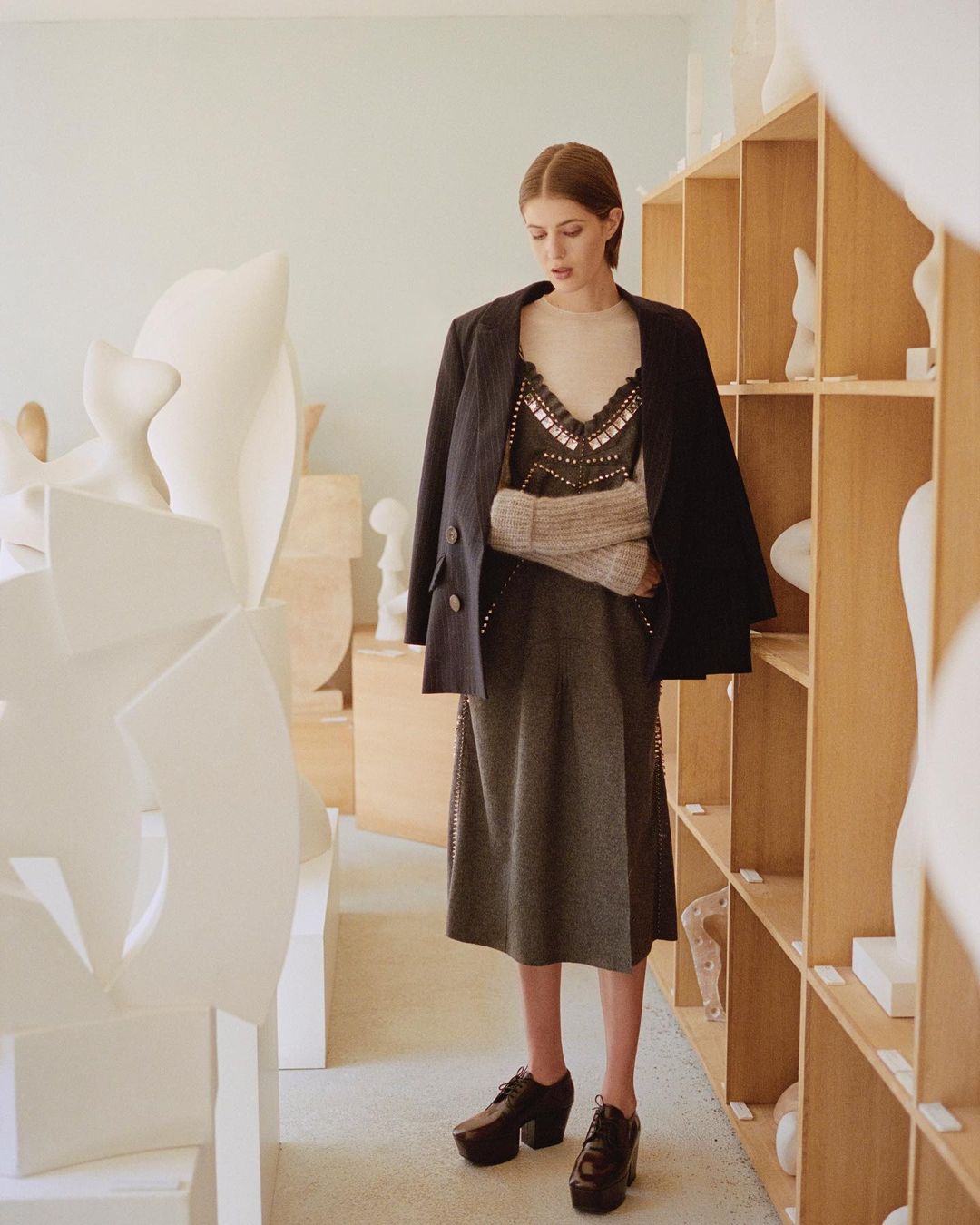 A woman in a blazer stands in an art studio surrounded by sculptures. She is wearing a dress and platform shoes, looking down.