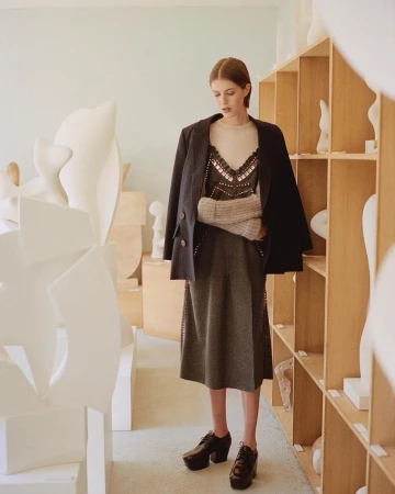 A woman in a blazer stands in an art studio surrounded by sculptures. She is wearing a dress and platform shoes, looking down.