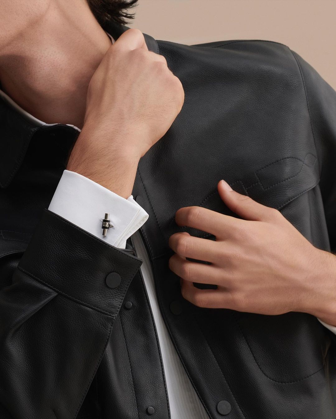 Adult male displaying elegant cufflinks and wearing a black leather jacket and a white dress shirt in a softly lit setting.