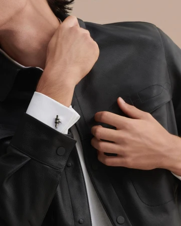 Adult male displaying elegant cufflinks and wearing a black leather jacket and a white dress shirt in a softly lit setting.