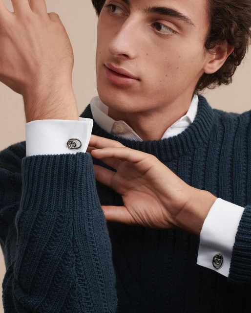 Close-up of a dark-haired, light-skinned adult male wearing a navy cable-knit sweater and shirt with cuff links on his forearms.