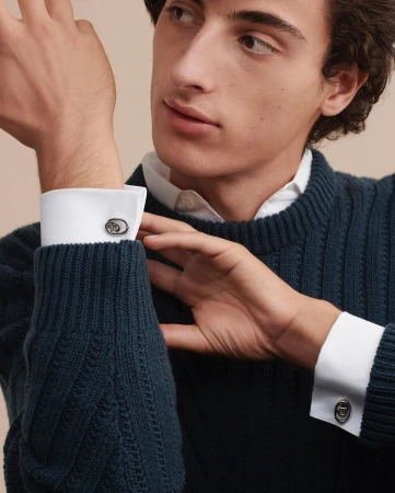 Close-up of a dark-haired, light-skinned adult male wearing a navy cable-knit sweater and shirt with cuff links on his forearms.