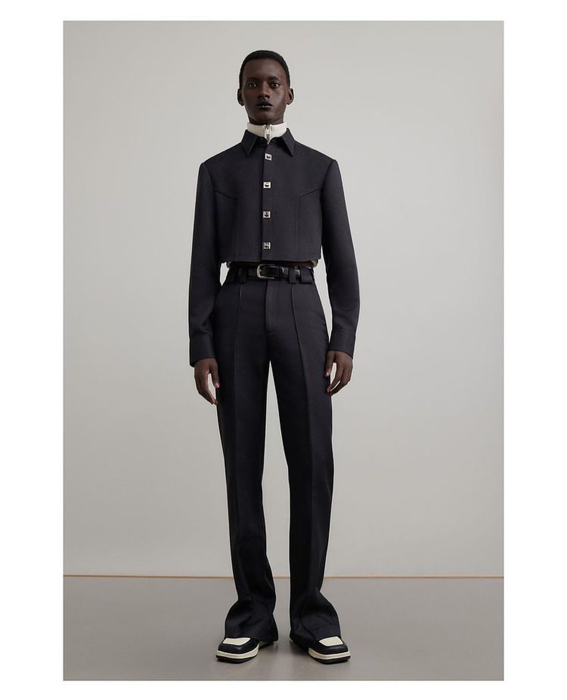 Dark-skinned male model in a black cropped shirt, dress pants, and black-and-white shoes poses in a studio setting.