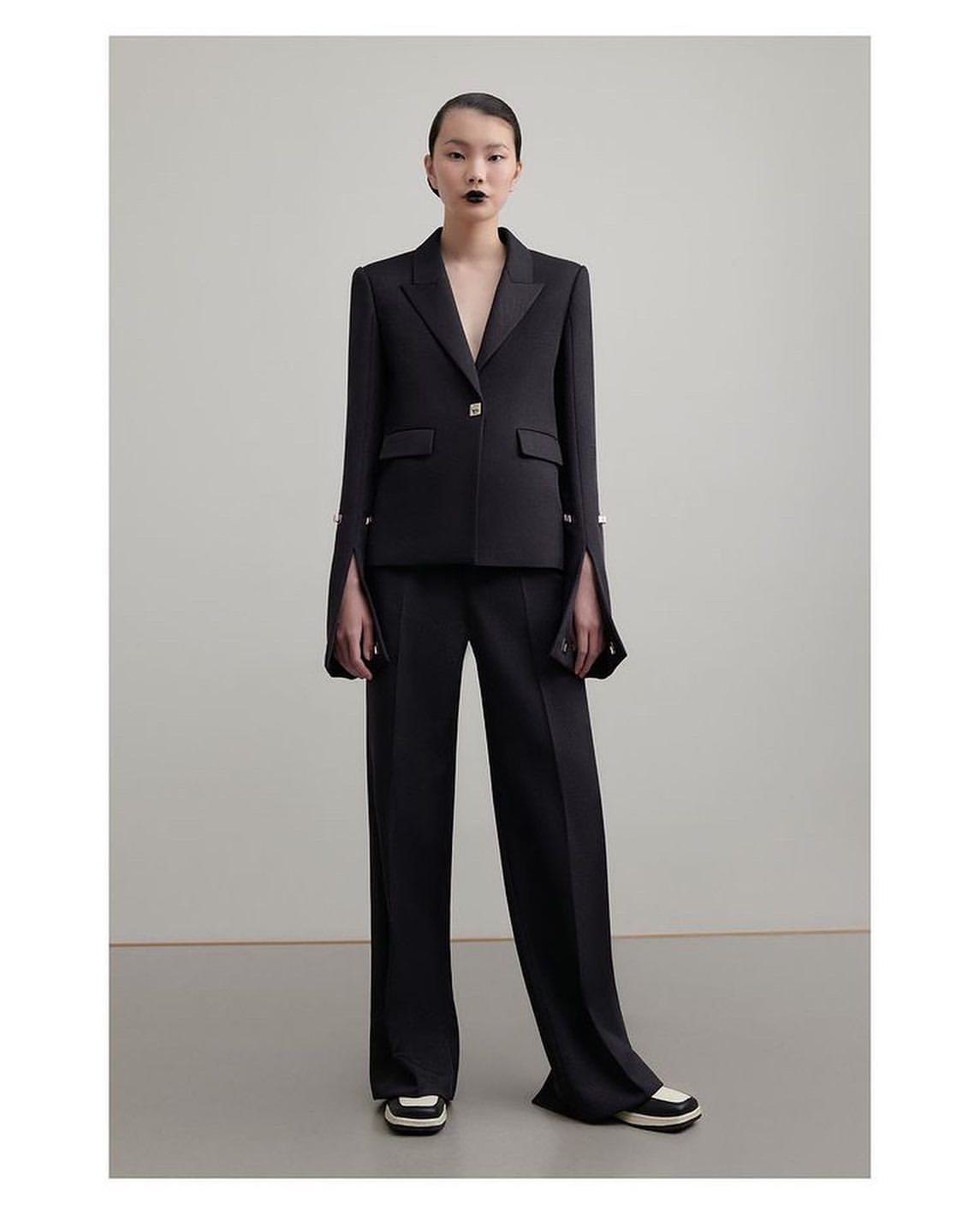 Model wearing a dark tailored suit with flared trousers and contrasting shoes against a neutral background