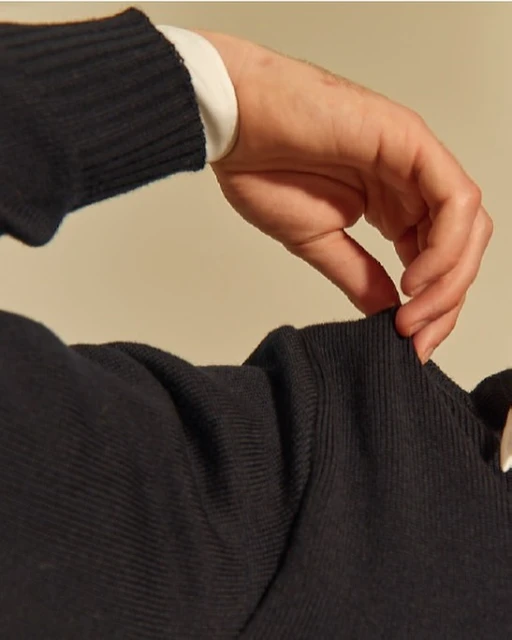 Close-up of a hand adjusting the sleeve of a dark ribbed sweater, revealing a white shirt cuff underneath, against a neutral background.