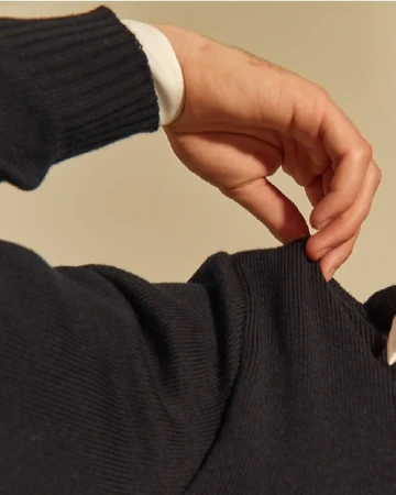 Close-up of a hand adjusting the sleeve of a dark ribbed sweater, revealing a white shirt cuff underneath, against a neutral background.