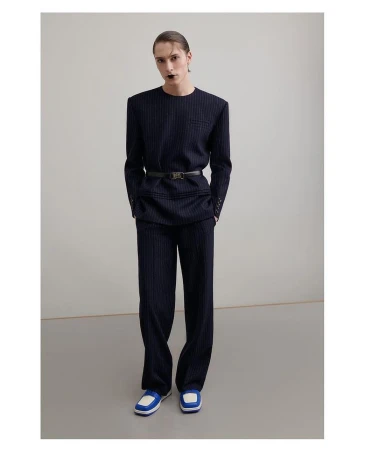 Full-length portrait of a slender adult wearing a pinstripe suit, cinched with a belt, and colorful athletic shoes in a studio.