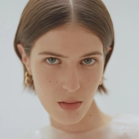 Close-up portrait of a woman with short parted brown hair and gold hoop earrings against a neutral background.