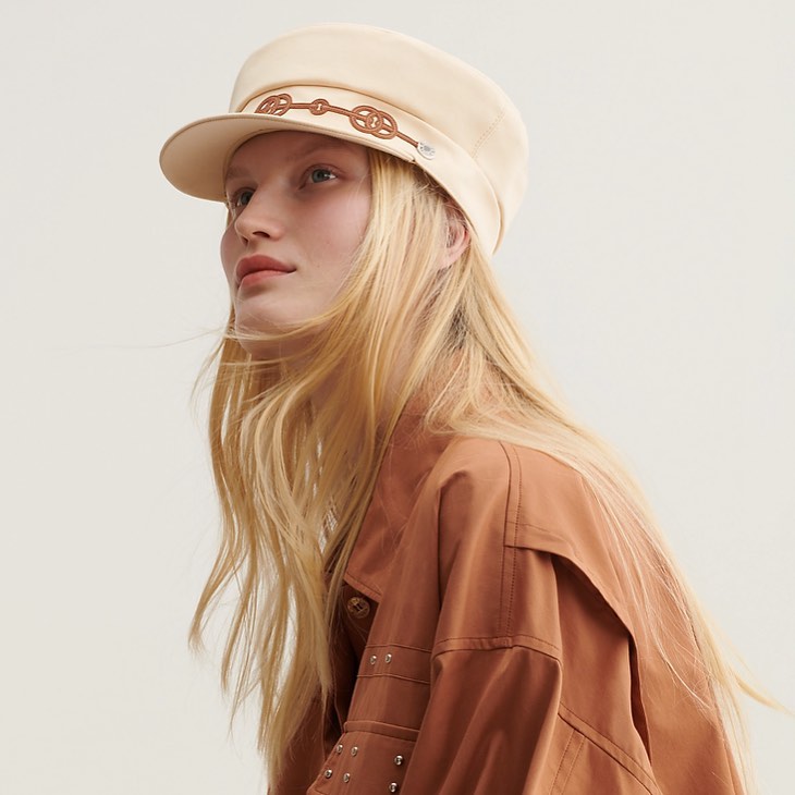 Woman in beige cap and brown jacket poses against a bright background in a studio shot, showcasing fashion and style.