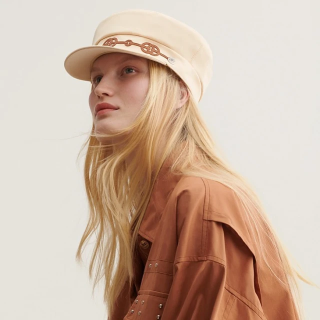 Woman in beige cap and brown jacket poses against a bright background in a studio shot, showcasing fashion and style.