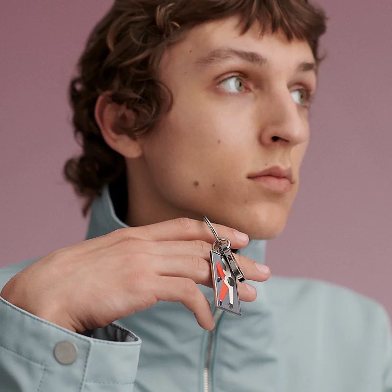 Young adult male with curly brown hair holds a modern, graphic key chain in front of a solid light purple background.