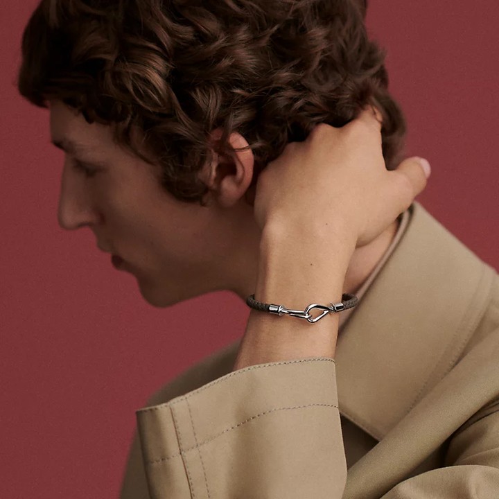 Man wearing a tan jacket and a grey bracelet with a silver clasp in front of a solid red background in a close-up studio shot.