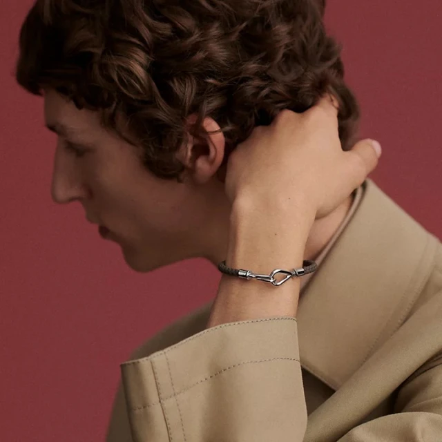Man wearing a tan jacket and a grey bracelet with a silver clasp in front of a solid red background in a close-up studio shot.