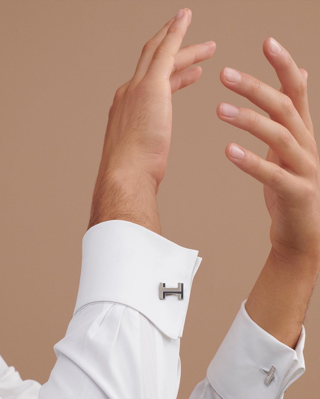 Close-up studio shot of a male hand displaying designer cufflinks on a crisp white dress shirt with a light brown backdrop.