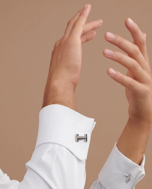 Close-up studio shot of a male hand displaying designer cufflinks on a crisp white dress shirt with a light brown backdrop.