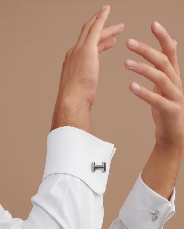Close-up studio shot of a male hand displaying designer cufflinks on a crisp white dress shirt with a light brown backdrop.