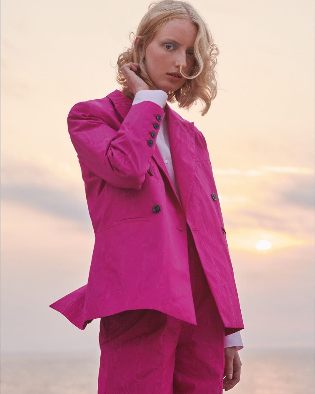 Woman in a pink suit poses outdoors against a sunset sky. She has blond hair and is holding her hand up to her neck.
