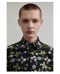 Portrait of a man with short hair wearing a black shirt embroidered with colorful floral patterns against a plain background