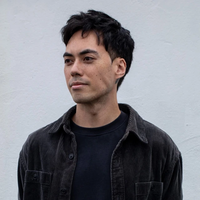 Portrait of a young adult man with dark hair, wearing a corduroy shirt, against a plain white wall in natural light, looking to the side.