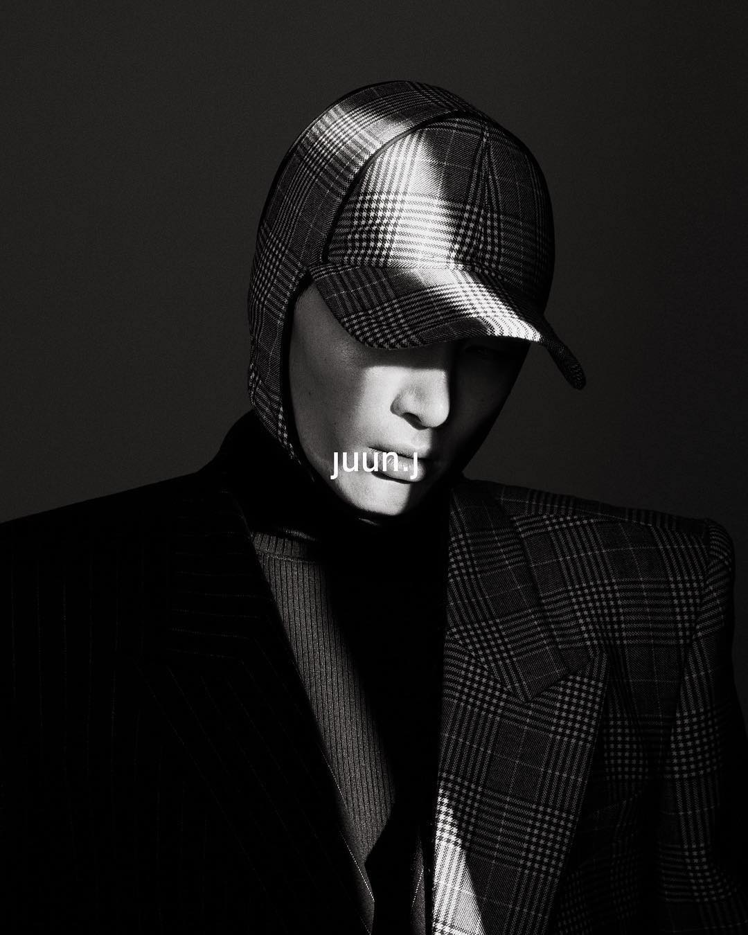 Monochrome portrait of a person wearing a plaid cap and blazer, highlighted with a beam of light against a dark background.