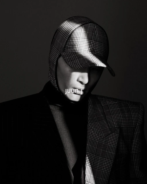 Monochrome portrait of a person wearing a plaid cap and blazer, highlighted with a beam of light against a dark background.