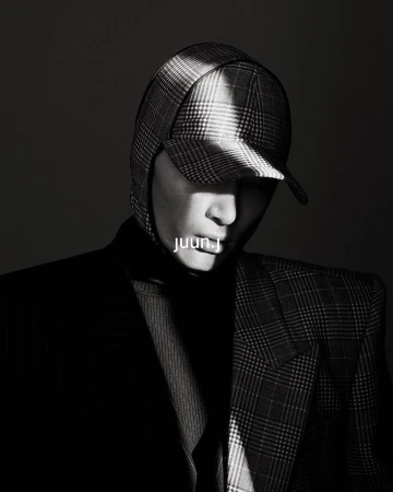 Monochrome portrait of a person wearing a plaid cap and blazer, highlighted with a beam of light against a dark background.