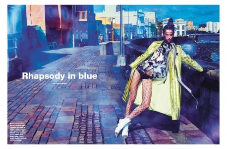 Editorial fashion shot of a fashion model in an iridescent coat and fishnets posing on a brick street with colorful buildings.