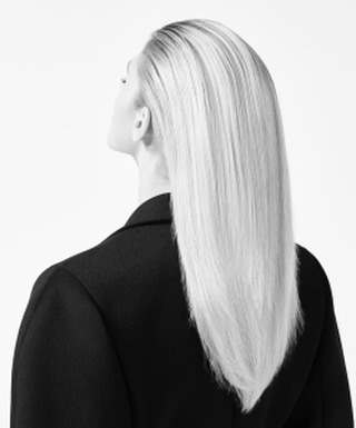 Back view of a blonde woman with long, straight hair wearing a black jacket against a bright white background in a studio setting.