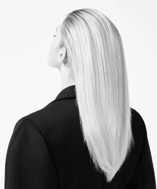 Back view of a blonde woman with long, straight hair wearing a black jacket against a bright white background in a studio setting.