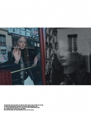 A young woman gazes out of a window in a Parisian street, reflecting the monochrome image of another woman in a melancholic scene.