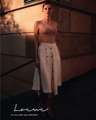 A woman poses in a tan top and cream skirt, standing next to a concrete wall with a sign, captured in soft, warm lighting.