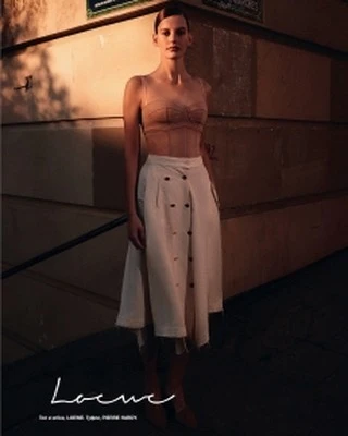 A woman poses in a tan top and cream skirt, standing next to a concrete wall with a sign, captured in soft, warm lighting.