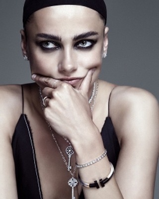 Close-up portrait of a woman with dramatic eye makeup, wearing layered jewelry, and a black top against a neutral gray backdrop.