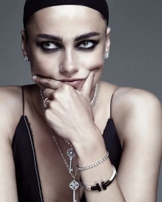Close-up portrait of a woman with dramatic eye makeup, wearing layered jewelry, and a black top against a neutral gray backdrop.
