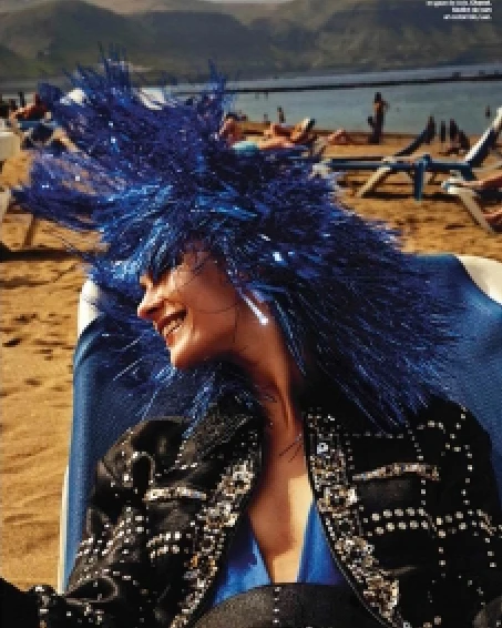Woman with a festive blue tinsel wig and embellished jacket smiles on a beach near blue deck chairs and sunbathers in the background.