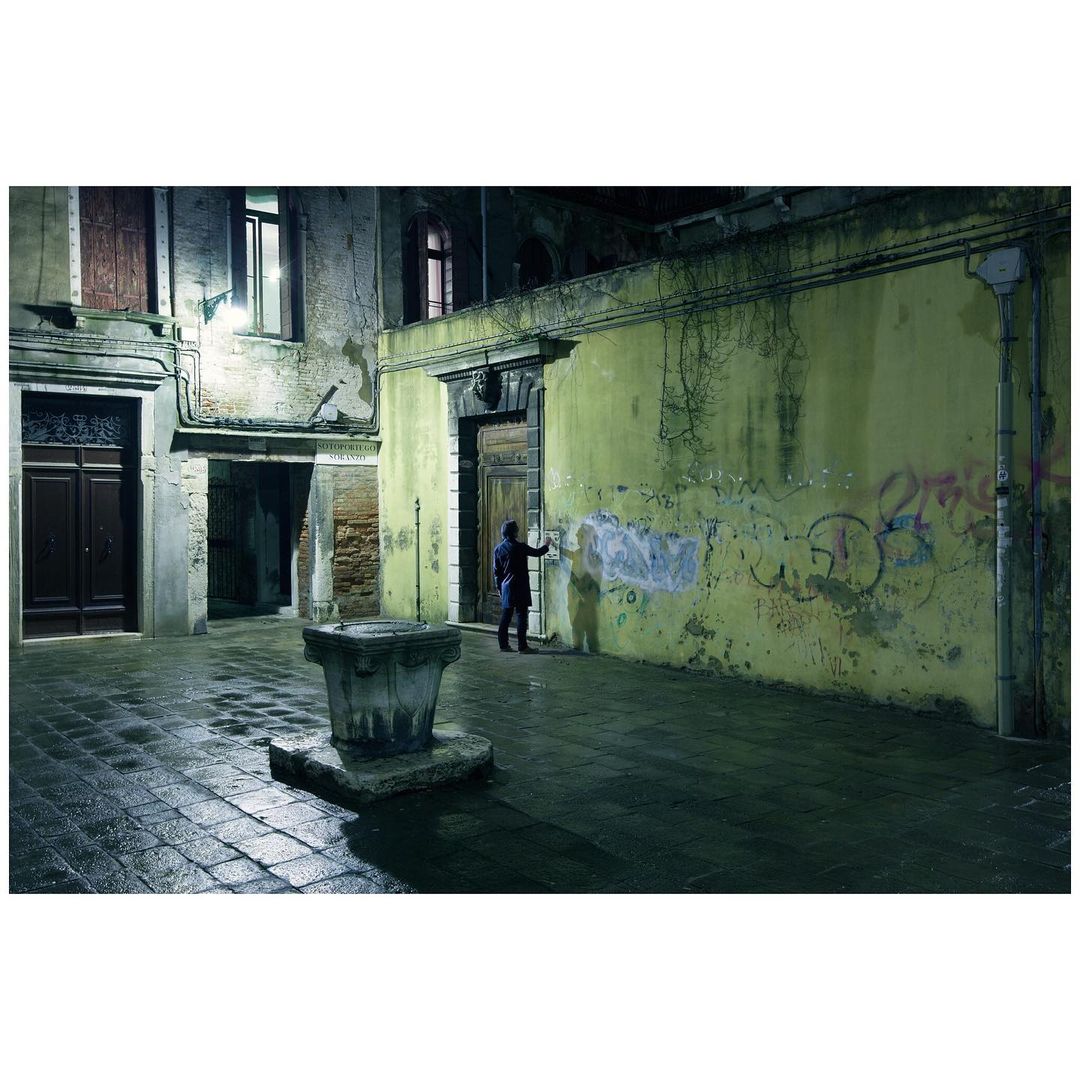Person in dark jacket painting graffiti on a weathered wall in a dimly lit, old European urban environment at night in Venice.