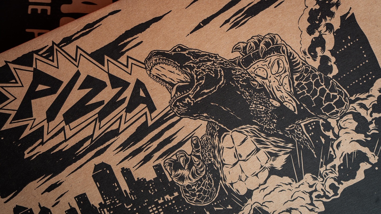 Graphic black and tan illustration on a pizza box features a monster eating pizza over a silhouetted cityscape with bold 'PIZZA' text.