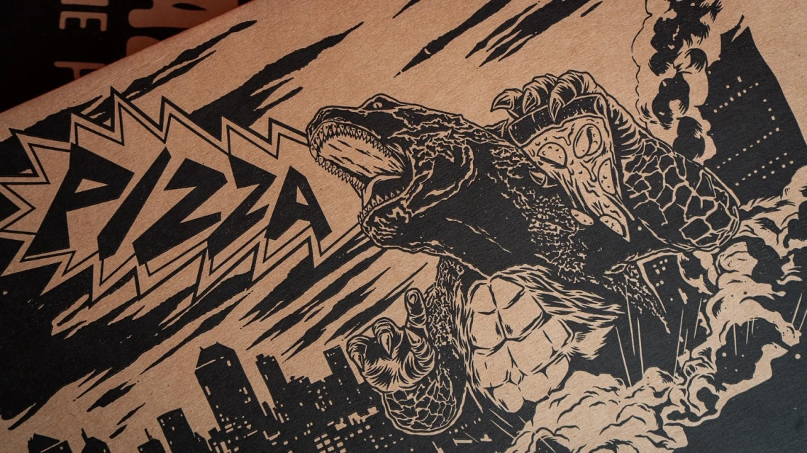 Graphic black and tan illustration on a pizza box features a monster eating pizza over a silhouetted cityscape with bold 'PIZZA' text.