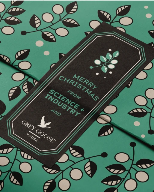 Close-up of a black and green Grey Goose Vodka promotional tag and background. The tag says 'Merry Christmas from Science + Industry'.