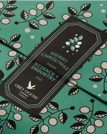 Close-up of a black and green Grey Goose Vodka promotional tag and background. The tag says 'Merry Christmas from Science + Industry'.