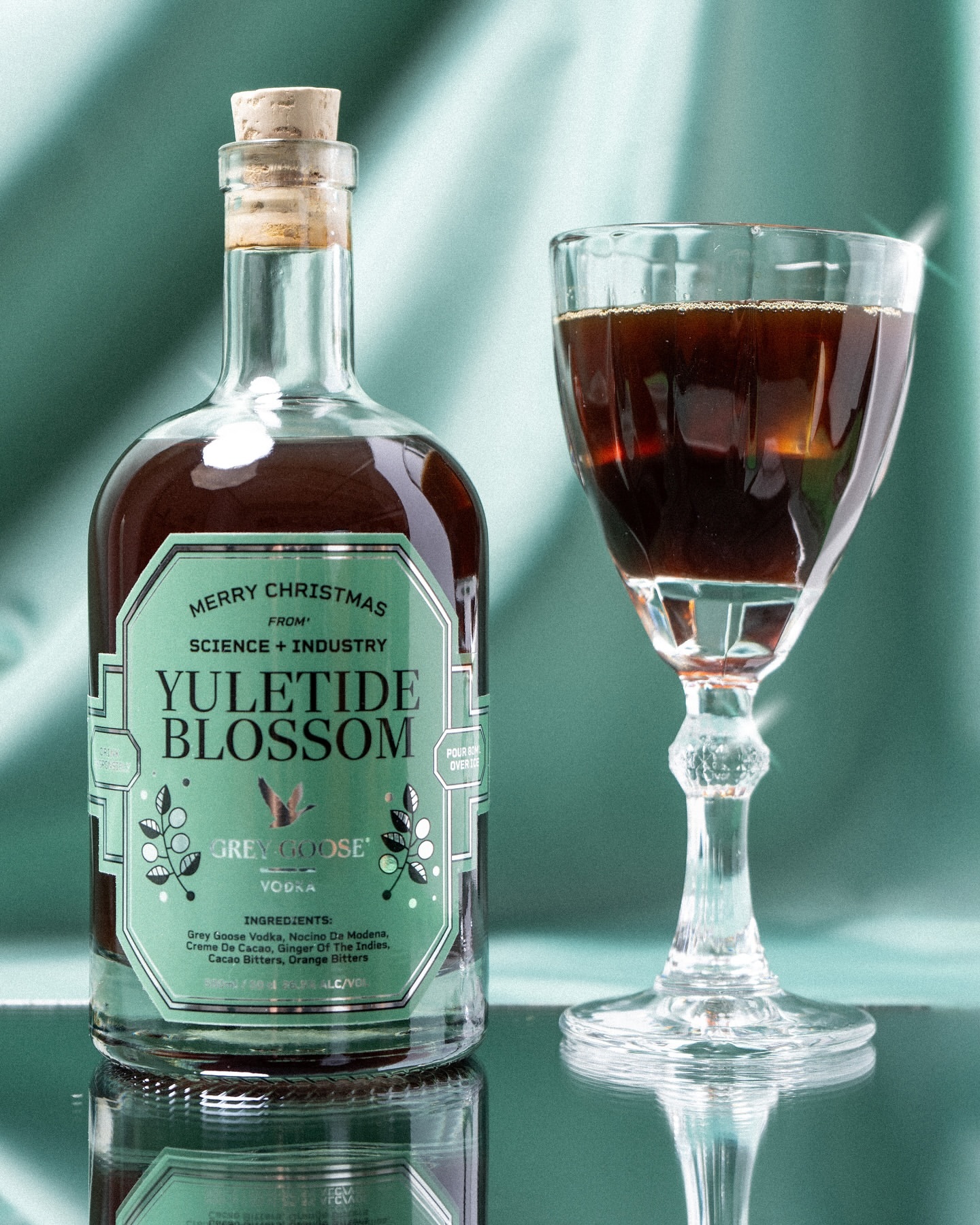 Vodka bottle next to an elegant cocktail glass filled with a dark drink against a teal, draped fabric background with surface reflections.