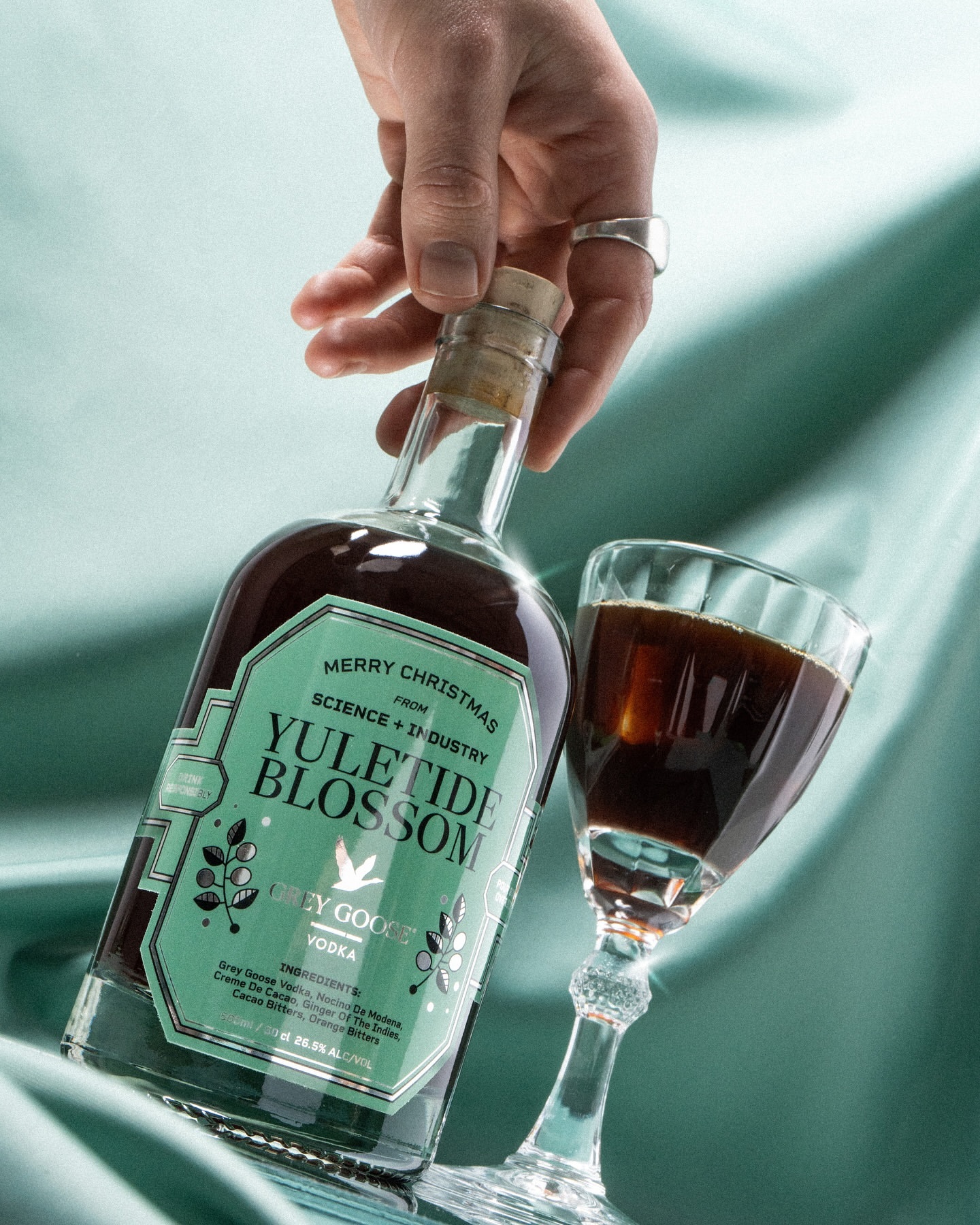 A person's hand holding a bottle of Yuletide Blossom Grey Goose vodka next to a stemmed glass filled with a dark liquid on blue fabric.