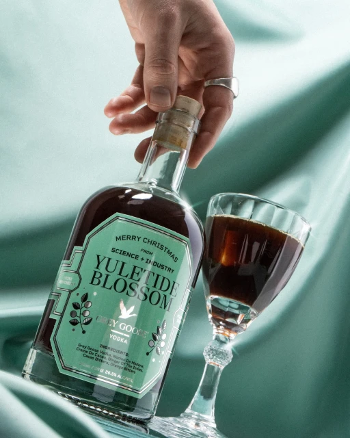 A person's hand holding a bottle of Yuletide Blossom Grey Goose vodka next to a stemmed glass filled with a dark liquid on blue fabric.
