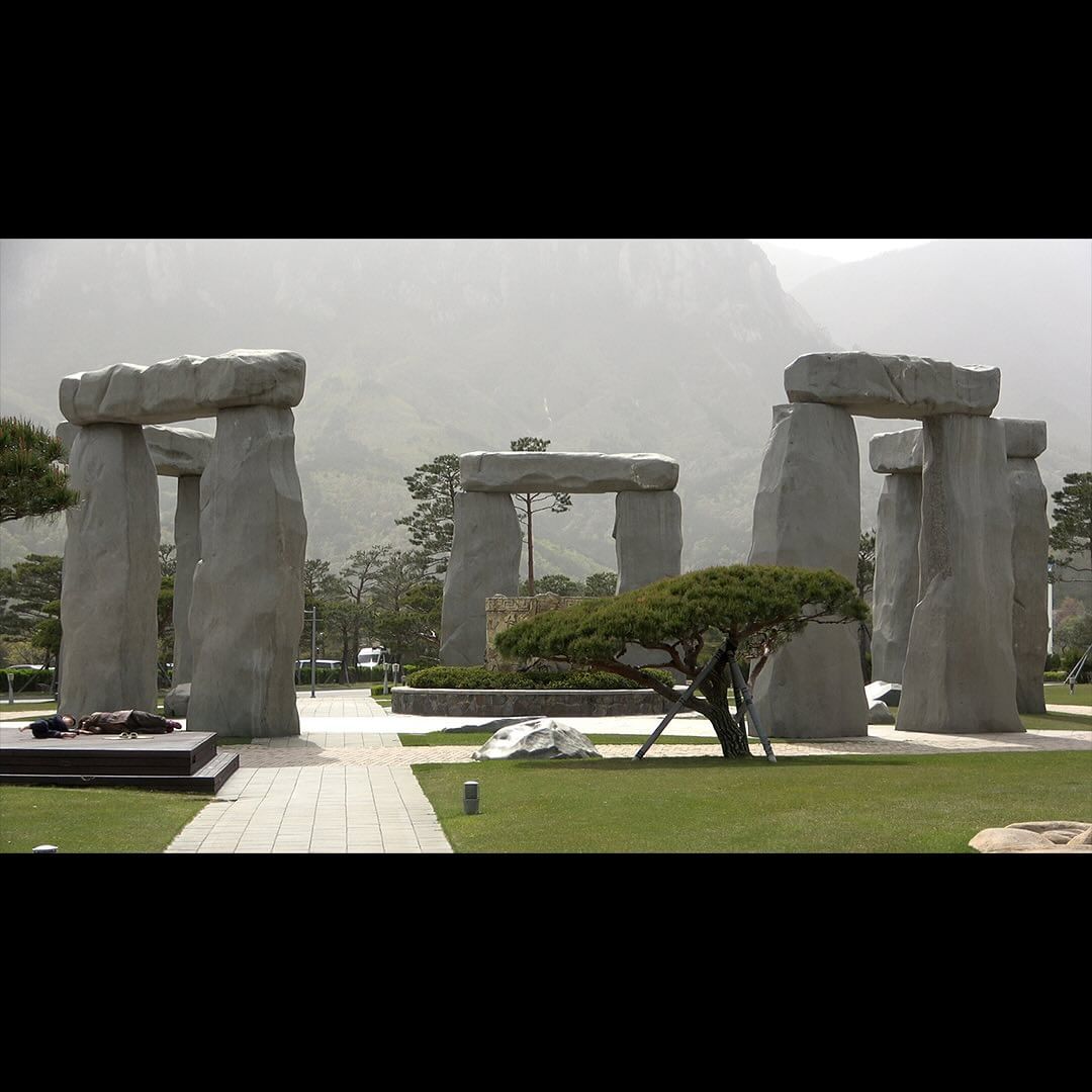Stonehenge replica stands on a green lawn with mountains visible in the background beneath a cloudy sky on an overcast day.