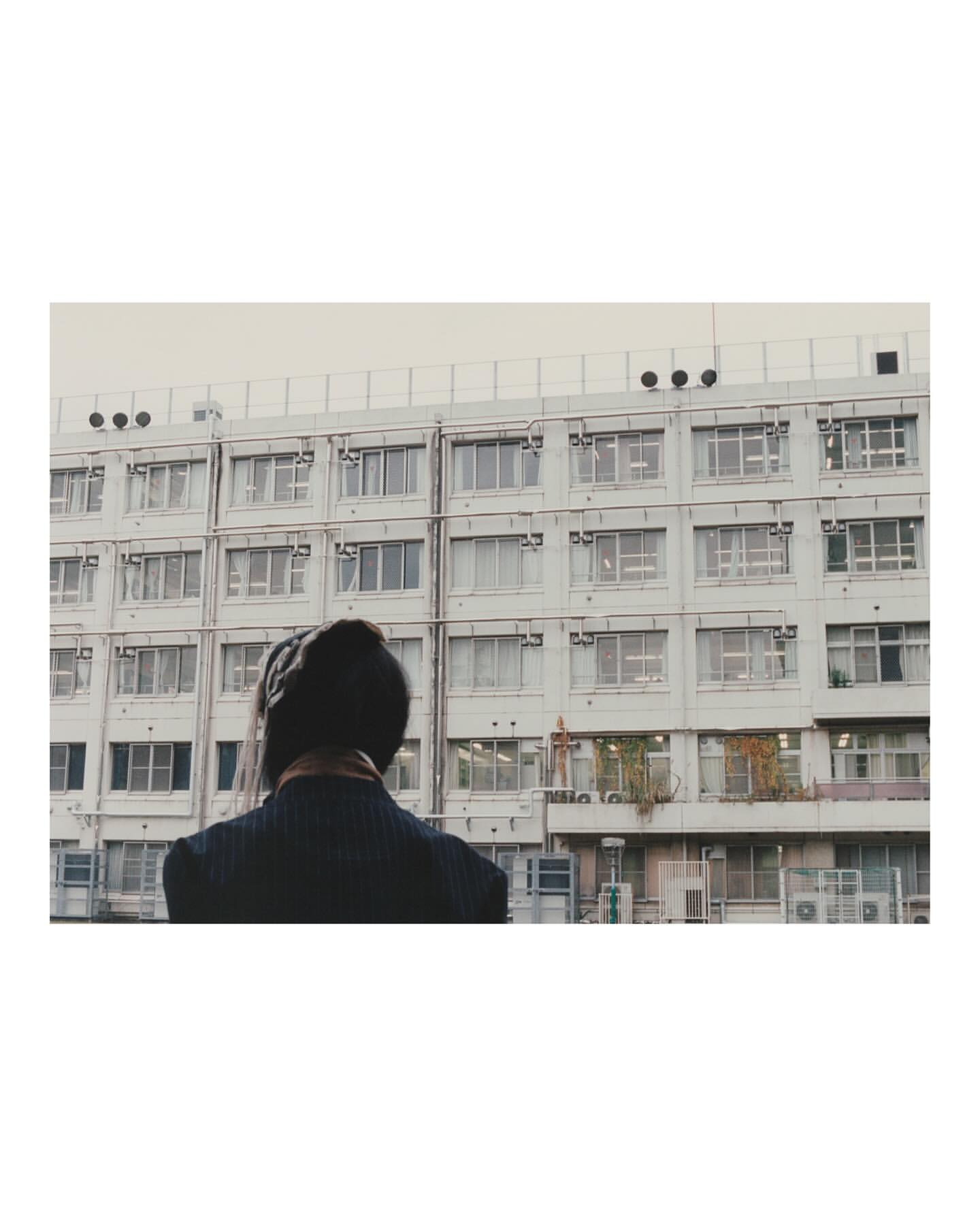 A woman faces a large concrete building with many windows, under a cloudy sky, creating an urban scene with neutral tones.