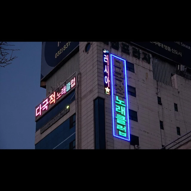 Exterior shot of a multi-story building with Korean text signs illuminated in neon lights against a dusk sky in an urban environment.