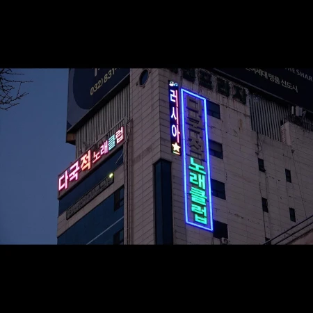 Exterior shot of a multi-story building with Korean text signs illuminated in neon lights against a dusk sky in an urban environment.